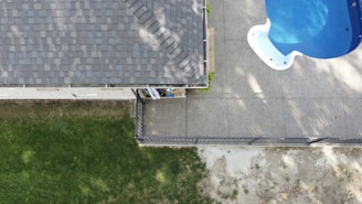 An aerial view captures a section of a house with a tiled roof on the left side and a backyard area. A concrete patio features a blue swimming pool in the upper right corner. The bottom left shows a grassy yard, and a narrow garden bed runs alongside the house.
