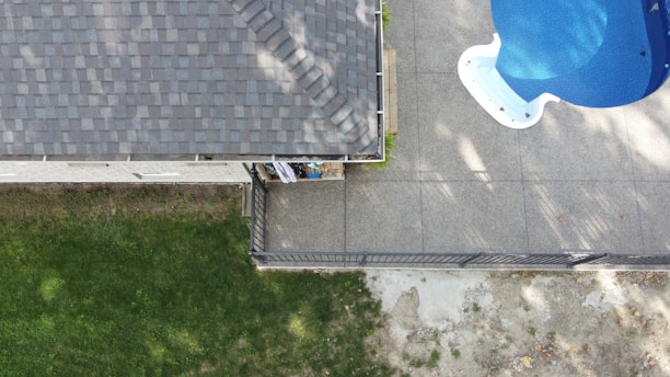 An aerial view captures a section of a house with a tiled roof on the left side and a backyard area. A concrete patio features a blue swimming pool in the upper right corner. The bottom left shows a grassy yard, and a narrow garden bed runs alongside the house.