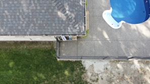 An aerial view captures a section of a house with a tiled roof on the left side and a backyard area. A concrete patio features a blue swimming pool in the upper right corner. The bottom left shows a grassy yard, and a narrow garden bed runs alongside the house.