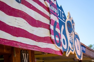 A Route 66 sign is prominently displayed with vintage styling. It is painted in bold colors and features an American flag motif. The sign is attached to a wooden structure. The blue sky adds a bright and outdoor atmosphere.