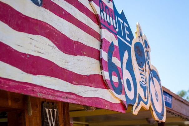 A Route 66 sign is prominently displayed with vintage styling. It is painted in bold colors and features an American flag motif. The sign is attached to a wooden structure. The blue sky adds a bright and outdoor atmosphere.