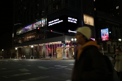 A nighttime urban scene features a modern building with colorful interior lighting. A large digital sign displays text messages. Two people are visible on the street, one walking and another using a phone. Bright streetlights illuminate the area, highlighting traffic signals and an advertisement for an event dated February 9.