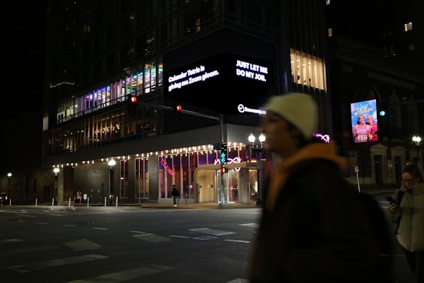 A nighttime urban scene features a modern building with colorful interior lighting. A large digital sign displays text messages. Two people are visible on the street, one walking and another using a phone. Bright streetlights illuminate the area, highlighting traffic signals and an advertisement for an event dated February 9.