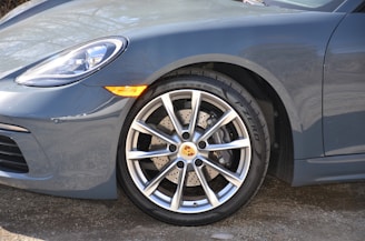 Close-up of the front wheel and headlight of a sleek, modern sports car with a metallic finish. The wheel features a high-performance brake system and an alloy rim with a prominently displayed logo.