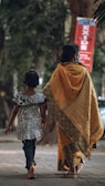 A woman and a child walk down a tree-lined street. The woman is wearing a traditional sari with a gold and orange pattern, while the child is in a black and white patterned dress paired with jeans. They both appear to be barefoot. In the background, there is a red sign with writing and lush green foliage.