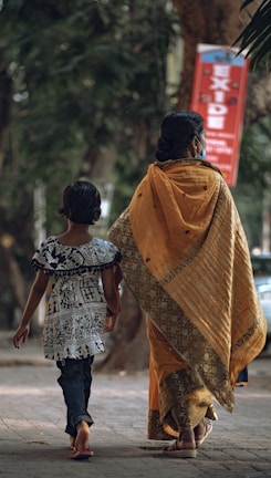 A woman and a child walk down a tree-lined street. The woman is wearing a traditional sari with a gold and orange pattern, while the child is in a black and white patterned dress paired with jeans. They both appear to be barefoot. In the background, there is a red sign with writing and lush green foliage.