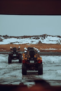 Two all-terrain vehicles are parked in a snowy and muddy area, surrounded by a rugged landscape with patches of snow and brown earth. The ATVs are aligned in a straight line, facing a rocky backdrop.