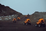A group of individuals wearing bright orange jackets and helmets are riding all-terrain vehicles up a rugged, rocky path. The terrain is barren with patches of snow scattered around, set against a cloudy sky.