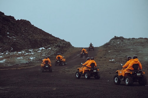 A group of individuals wearing bright orange jackets and helmets are riding all-terrain vehicles up a rugged, rocky path. The terrain is barren with patches of snow scattered around, set against a cloudy sky.
