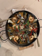 A delicious seafood paella with vibrant ingredients including large prawns, mussels, clams, peas, and red bell peppers arranged over a bed of saffron-infused rice in a traditional round paella pan. The dish is garnished with lemon wedges and fresh herbs, providing a colorful and appetizing presentation.