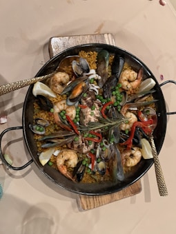A delicious seafood paella with vibrant ingredients including large prawns, mussels, clams, peas, and red bell peppers arranged over a bed of saffron-infused rice in a traditional round paella pan. The dish is garnished with lemon wedges and fresh herbs, providing a colorful and appetizing presentation.