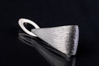 A sleek, silver metallic pendant with a textured surface lying on a black reflective surface. The pendant has an elongated triangular shape with a looped holder at one end.