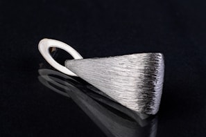 A sleek, silver metallic pendant with a textured surface lying on a black reflective surface. The pendant has an elongated triangular shape with a looped holder at one end.