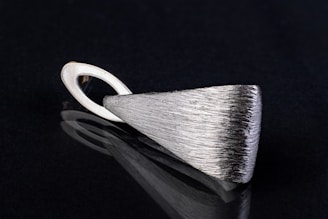 A sleek, silver metallic pendant with a textured surface lying on a black reflective surface. The pendant has an elongated triangular shape with a looped holder at one end.