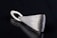 A sleek, silver metallic pendant with a textured surface lying on a black reflective surface. The pendant has an elongated triangular shape with a looped holder at one end.