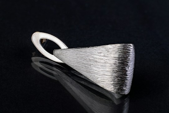 A sleek, silver metallic pendant with a textured surface lying on a black reflective surface. The pendant has an elongated triangular shape with a looped holder at one end.