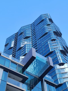A modern high-rise building with a unique architectural design featuring wavy lines and extensive use of glass. A striking glass-bottom swimming pool extends outward from one of the upper floors, adding an element of luxury and innovation. The building facade reflects the clear blue sky, enhancing its contemporary and sleek appearance.