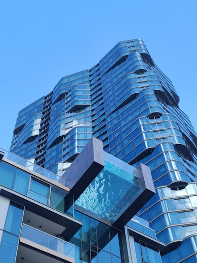 A modern high-rise building with a unique architectural design featuring wavy lines and extensive use of glass. A striking glass-bottom swimming pool extends outward from one of the upper floors, adding an element of luxury and innovation. The building facade reflects the clear blue sky, enhancing its contemporary and sleek appearance.