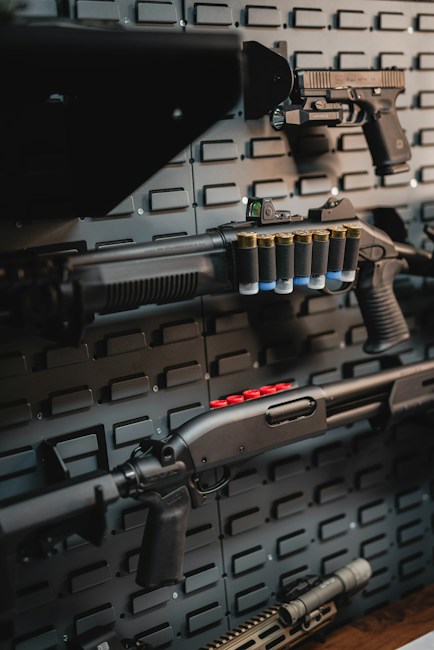 A collection of firearms is mounted on a dark, textured wall. The arrangement includes various types of rifles and handguns, each equipped with different tactical accessories and attachments. Shells are visible on some of the firearms, contributing to the overall military or tactical theme.