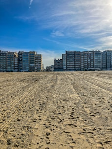 A sandy beach stretches into the distance with various footprints and tire tracks. Residential buildings line the horizon under a clear blue sky, with a prominent, turreted structure at the center.