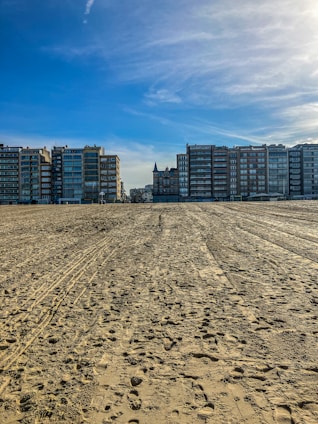 A sandy beach stretches into the distance with various footprints and tire tracks. Residential buildings line the horizon under a clear blue sky, with a prominent, turreted structure at the center.