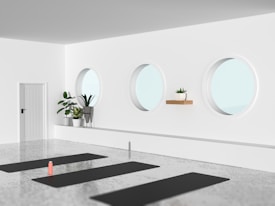 A minimalist room with three circular windows, several plants in white pots, a small wooden shelf, and yoga mats on a polished concrete floor. A water bottle is placed on one of the mats.