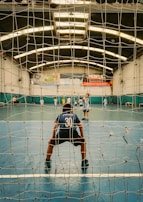 A young player, wearing a jersey with the number 30, stands in front of a goal net on an indoor sports court. Several other players are visible in the background, scattered across the court, engaged in a game, possibly soccer or futsal. The indoor space is well-lit with fluorescent lights, and banners hang on the walls.