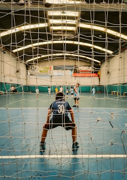 A young player, wearing a jersey with the number 30, stands in front of a goal net on an indoor sports court. Several other players are visible in the background, scattered across the court, engaged in a game, possibly soccer or futsal. The indoor space is well-lit with fluorescent lights, and banners hang on the walls.