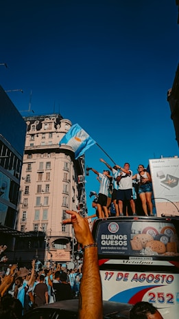 A group of people are celebrating on top of a bus in a city setting. They are waving flags and wearing matching sports jerseys, suggesting a sports victory celebration. The crowd below is engaged with raised arms and cheering. The background features historic architecture and advertisements.