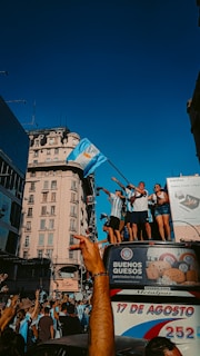 A group of people are celebrating on top of a bus in a city setting. They are waving flags and wearing matching sports jerseys, suggesting a sports victory celebration. The crowd below is engaged with raised arms and cheering. The background features historic architecture and advertisements.