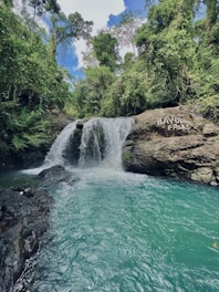 A serene waterfall cascades into a turquoise pool surrounded by lush, green foliage. The waterfall, known as Bayug Falls, is nestled in a dense forest with large rocks framing the scene. The sky is partly cloudy, creating a tranquil and inviting atmosphere.