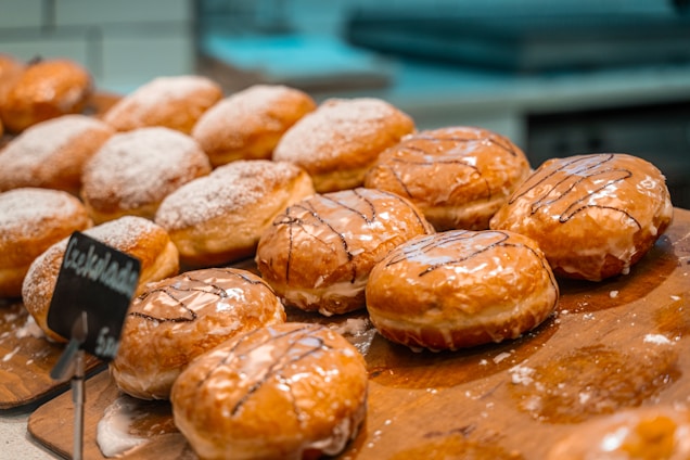 A selection of glazed donuts with chocolate drizzle is displayed on a wooden tray. Some donuts are dusted with powdered sugar, and a small chalkboard sign indicates the flavor. The background is slightly blurred, suggesting a bakery setting.