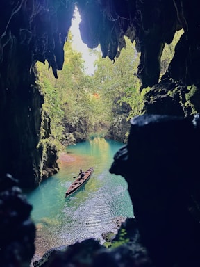 A serene turquoise river winds through a cavernous landscape, surrounded by lush greenery. A person paddles alone in a wooden canoe, adding a sense of scale and exploration. The cave's jagged edges frame the scene from above, creating a natural archway.