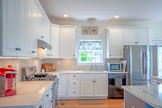 A modern kitchen with white cabinetry and stainless steel appliances. The room features a gas stove with overhead range hood, a large refrigerator, and a microwave. A coffee maker is situated on the counter, which has a smooth, light-colored surface. There's a window above the sink with a decorative blue and white curtain, and small decor items are placed on the counters.