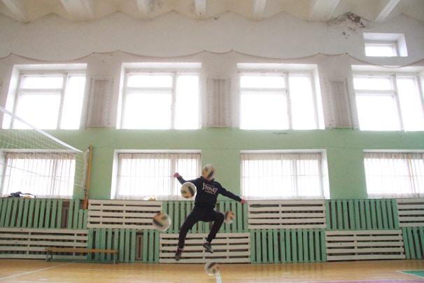 A person is jumping in an indoor gymnasium, surrounded by multiple volleyballs in mid-air. The gym has large windows, green walls with wooden paneling, and a volleyball net on one side. The floor is wooden, and there are light fixtures on the ceiling.