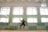 A person is jumping in an indoor gymnasium, surrounded by multiple volleyballs in mid-air. The gym has large windows, green walls with wooden paneling, and a volleyball net on one side. The floor is wooden, and there are light fixtures on the ceiling.