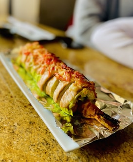 A dish presented on a rectangular white plate lined with foil, featuring a sushi roll wrapped in thin strips of meat. The roll is garnished with finely chopped red garnish and rests on a bed of green lettuce. The surrounding surface is a speckled, light brown countertop.