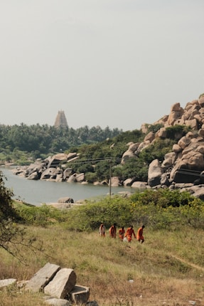 A serene landscape featuring a river surrounded by lush greenery and large boulders. In the distance, there is a visible temple tower amidst palm trees. A group of people dressed in orange garments walk along a grassy path in the foreground.