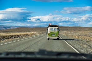 A vintage green van with a roof rack is driving on a long, straight road through a vast, open landscape. The sky is mostly clear with some clouds, and the road is bordered by dry, grassy plains extending to the horizon.