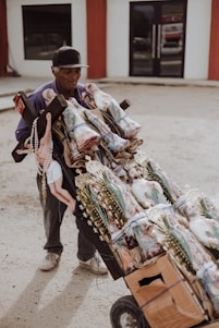 An elderly man is pushing a cart full of religious statues and items. The cart is made of cardboard and tied with blue string, holding several statues of religious figures, possibly saints, adorned with golden decorations and rosaries. The man is wearing a cap and appears to be in an outdoor setting with a building in the background.