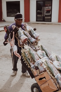 An elderly man is pushing a cart full of religious statues and items. The cart is made of cardboard and tied with blue string, holding several statues of religious figures, possibly saints, adorned with golden decorations and rosaries. The man is wearing a cap and appears to be in an outdoor setting with a building in the background.