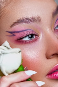 An up-close view of a person's face with artistic makeup, featuring vivid pink and purple eyeshadow and a geometric design extending from the eye. A white rose is held near the face, adding a touch of softness to the image.