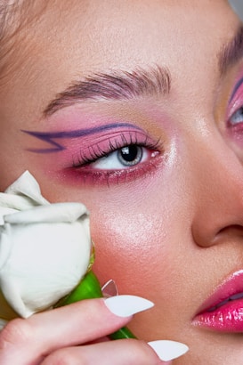 An up-close view of a person's face with artistic makeup, featuring vivid pink and purple eyeshadow and a geometric design extending from the eye. A white rose is held near the face, adding a touch of softness to the image.