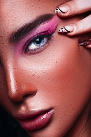 A close-up of a person's face featuring detailed eye makeup with bright pink eyeshadow. The eye is blue, and the eyebrows are dark and well-defined. The skin has visible freckles and a glossy finish. A hand with fingernails decorated with black designs is gently touching the eye area.