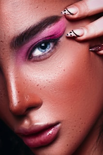 A close-up of a person's face featuring detailed eye makeup with bright pink eyeshadow. The eye is blue, and the eyebrows are dark and well-defined. The skin has visible freckles and a glossy finish. A hand with fingernails decorated with black designs is gently touching the eye area.