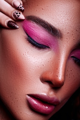 A close-up of a woman's face with vibrant pink eyeshadow and glossy pink lips. Her skin appears smooth and slightly freckled, and she has well-groomed eyebrows. A hand with artistically painted nails is touching her forehead.