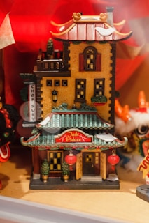 A small, intricately detailed model of a Chinese-style building named 'Jade Palace.' The structure features traditional architectural elements such as green tiled roofs with red accents and ornamental details. Red lanterns are hanging at the entrance, adding to the cultural theme. The walls are painted in a warm yellow color, and there are small potted plants placed on either side of the main entrance. The background includes decorative elements in bright colors, contributing to a festive atmosphere.