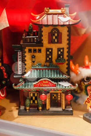 A small, intricately detailed model of a Chinese-style building named 'Jade Palace.' The structure features traditional architectural elements such as green tiled roofs with red accents and ornamental details. Red lanterns are hanging at the entrance, adding to the cultural theme. The walls are painted in a warm yellow color, and there are small potted plants placed on either side of the main entrance. The background includes decorative elements in bright colors, contributing to a festive atmosphere.