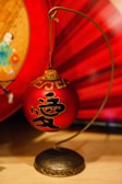 A decorative red ornament with black Chinese characters is suspended on a curved metal stand. The background features rich red hues, possibly from a fabric or paper, with the edge showing a partial illustration depicting a person in traditional attire.