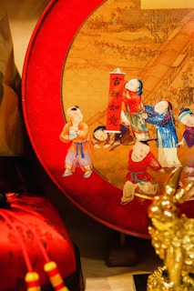 A decorative item with an intricate design depicting children dressed in traditional clothing, vividly painted, and set against a red circular background. Chinese calligraphy is prominently displayed on a vertical red strip. The surrounding elements include additional festive decorations and a golden sculpture.